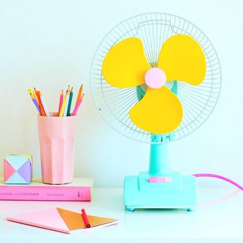 Photo d'un bureau avec la mise en scène d'un ventilateur bleu turquoise aux élices jaunes, d'un pot à crayon rose pastel, d'un livre rose et d'un bloc note rose et marron