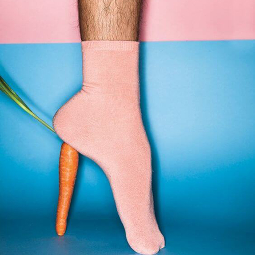 Jambe d'un homme portant une chaussette rose dont le talon est sur-élevé par une carotte représentant le talon d'une chaussure de femme